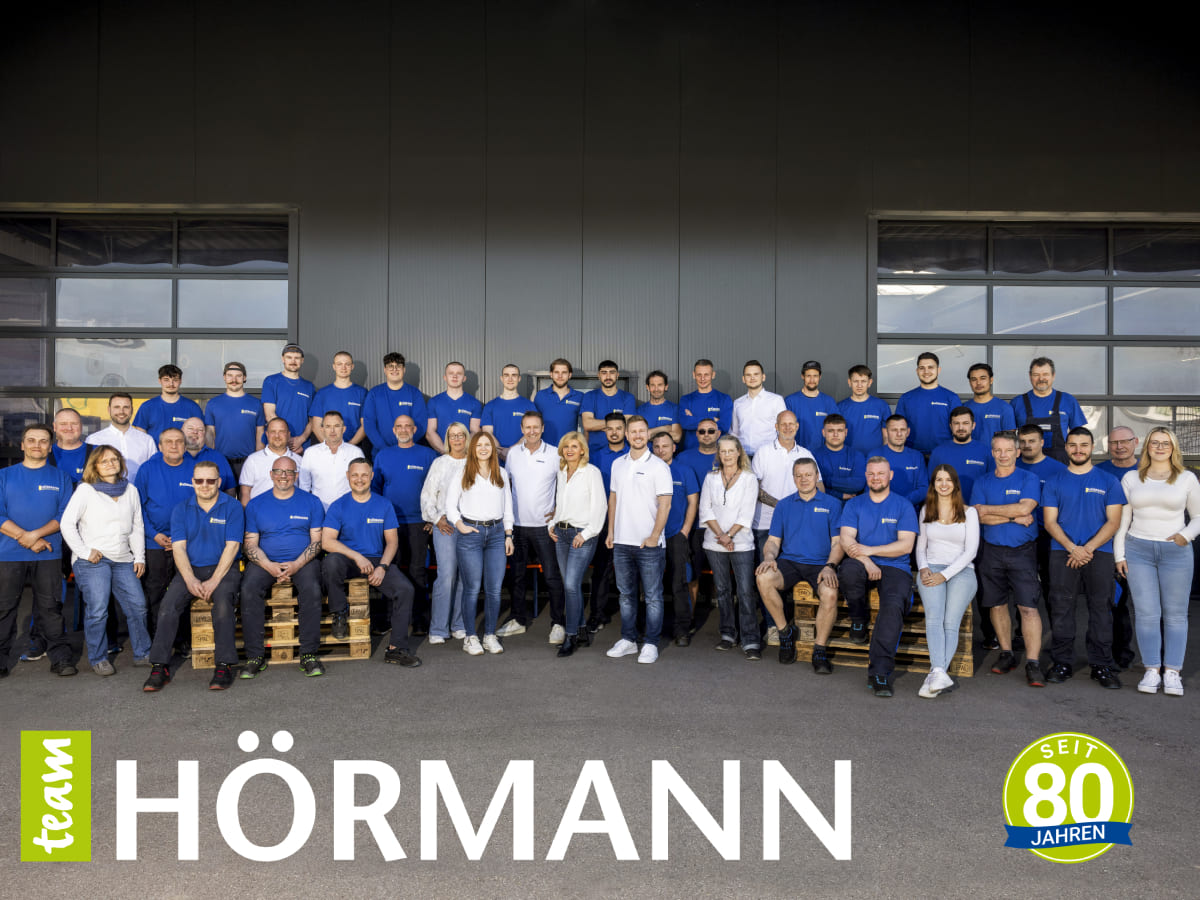 Hörmann Teamfoto zum 80-jährigen Jubiläum Großes Gruppenfoto von Hörmann-Mitarbeitenden in blauer und weißer Kleidung vor dem Firmengebäude mit 80-Jahre-Logo.