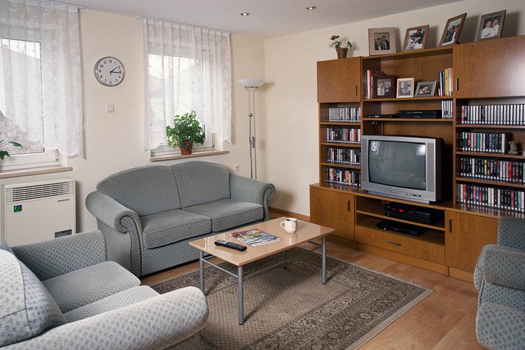 Ein Wohnzimmer mit grauer Couch und Schrankwand, in der Mitte ein Tisch auf einem Teppich.