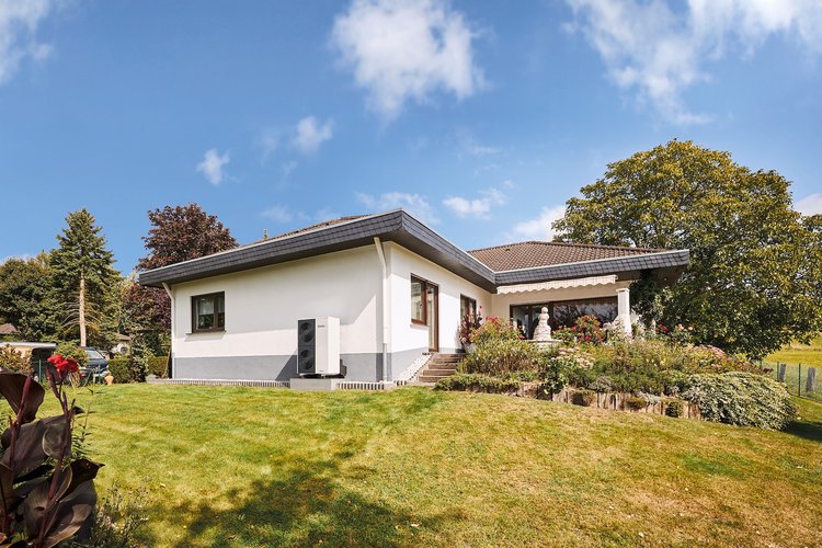 Ein Wohnhaus mit Vaillant-Wärmepumpe an der Hausfassade, im Vordergrund Garten und im Hintergrund blauer Himmel