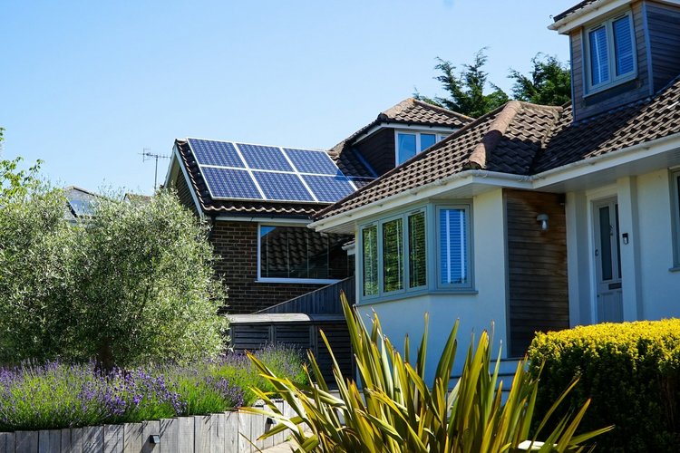Ein Wohnhaus mit PV-Anlage auf dem Dach, im Vordergrund Gärten, im Hintergrund blauer Himmel.
