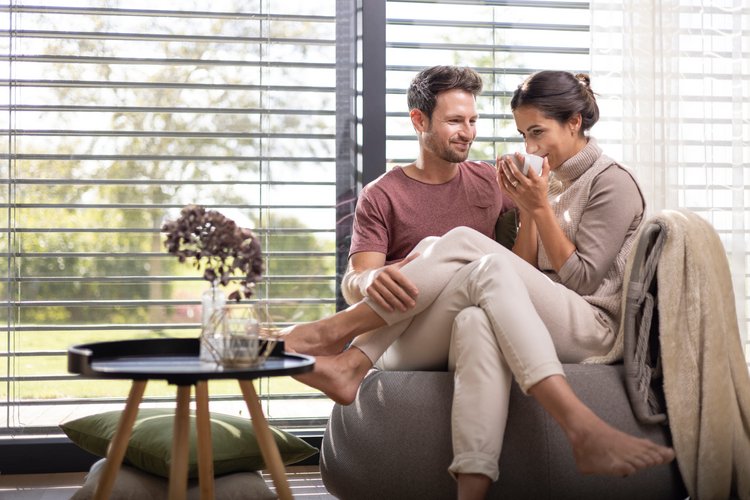 Paar sitzt barfuß und entspannt auf einem Sofa vor bodentiefem Fenster mit Lamellen, trinkt gemeinsam Kaffee.