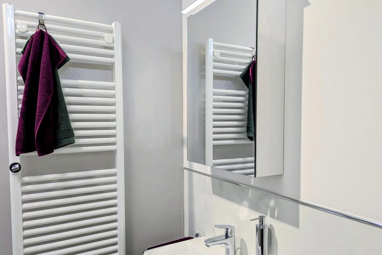 Modernes Badezimmer mit weißem Handtuchheizkörper, daran hängende Kleidung, Spiegel und Waschtisch im Vordergrund.