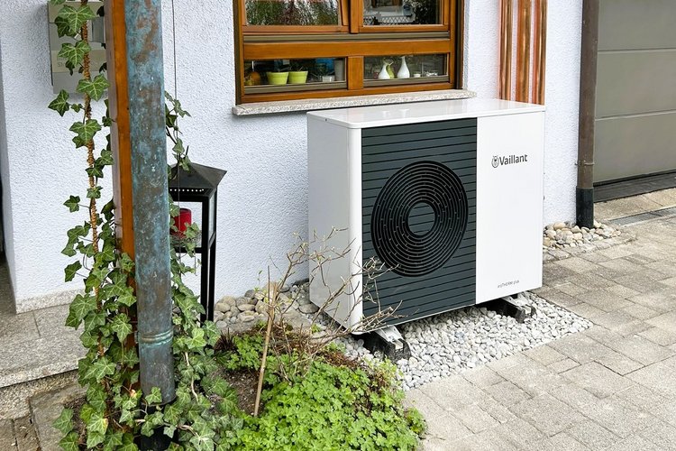 Wärmepumpen-Außeneinheit neben einem Fenster mit Holzrahmen an weißer Hauswand, umgeben von Deko-Elementen und Pflanzen.