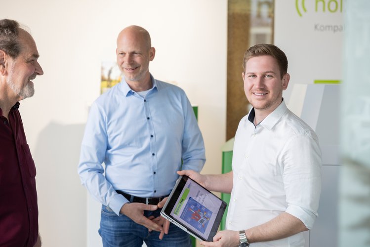 Drei Männer stehen in einem hellen Büro, einer zeigt ein Tablet mit farbiger Darstellung und alle lachen freundlich.