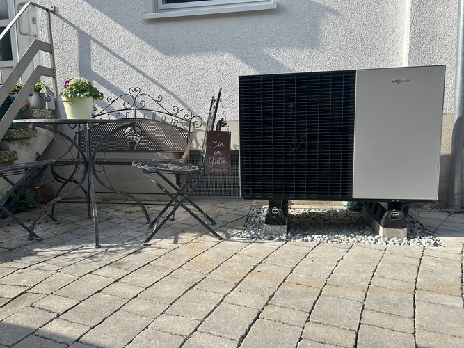 Wärmepumpen-Außeneinheit auf gepflasterter Terrasse neben Metallgartenmöbeln vor einer weiß verputzten Hauswand installiert.