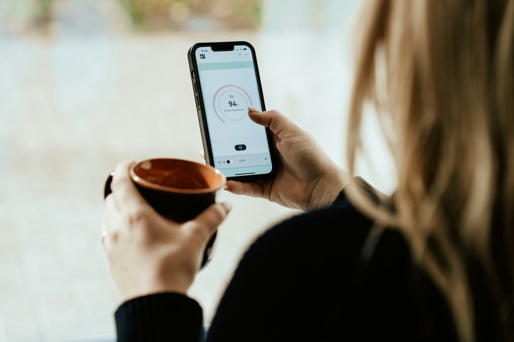 Frau hält Kaffeetasse in der einen Hand und Smartphone mit geöffneter Heizung-App in der anderen.