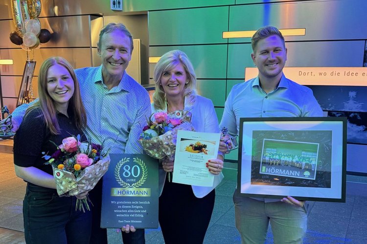 Vier lächelnde Personen in festlicher Umgebung mit Geschenken, Blumen und einem gerahmten Poster bei 80. Jubiläumsfeier.